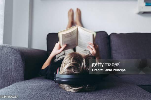 boy listening music while reading book on sofa at home - reading fotografías e imágenes de stock
