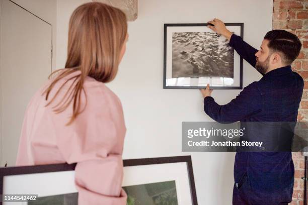 male and female are hanging a picture frame up on the white wall. - house for an art lover stock pictures, royalty-free photos & images