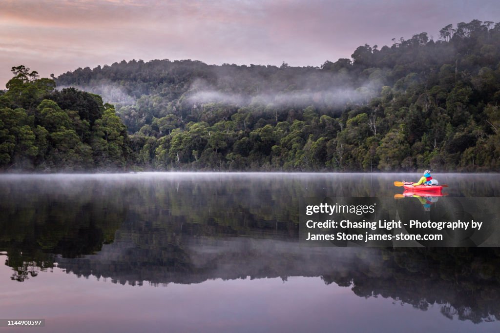 A girl enjoys a tranquil paddle in a kayak at dawn down a flat river with reflections of misty rainforest and mountain scenery
