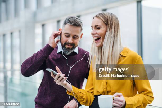 two businesspeople with a cup of coffee in a modern office, using smartphone and earphones. - office coffee break stock pictures, royalty-free photos & images