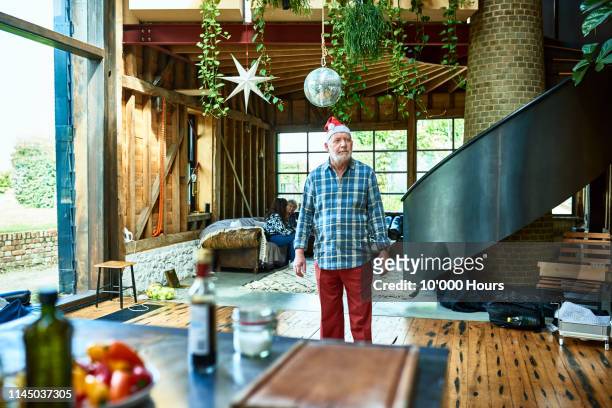 senior man wearing santa hat standing in middle of room - grumpy old man stock pictures, royalty-free photos & images