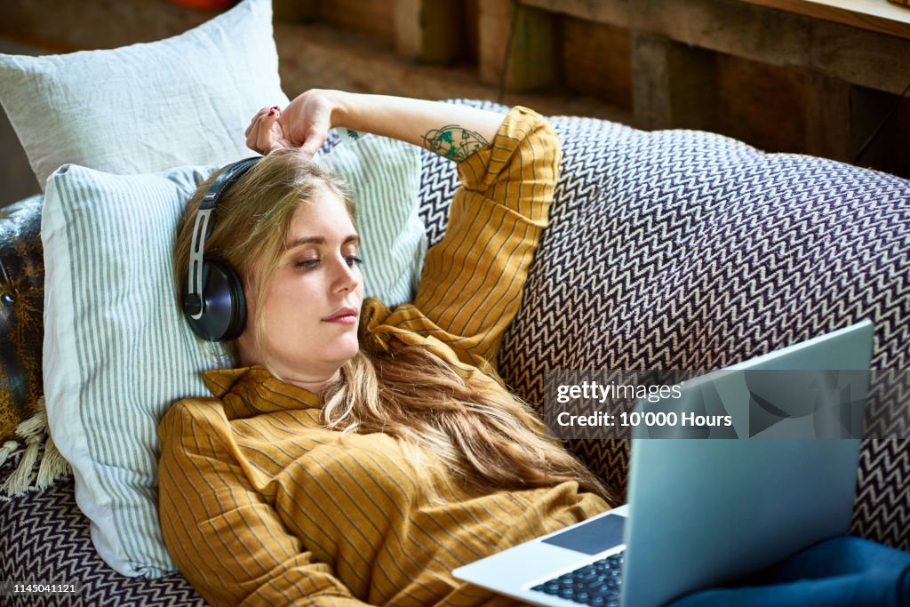 Mid adult woman reclining on sofa with laptop wearing headphones