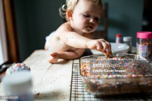 toddler girl sneaking colorful sprinkles off of freshly baked cake - messy cake stock pictures, royalty-free photos & images