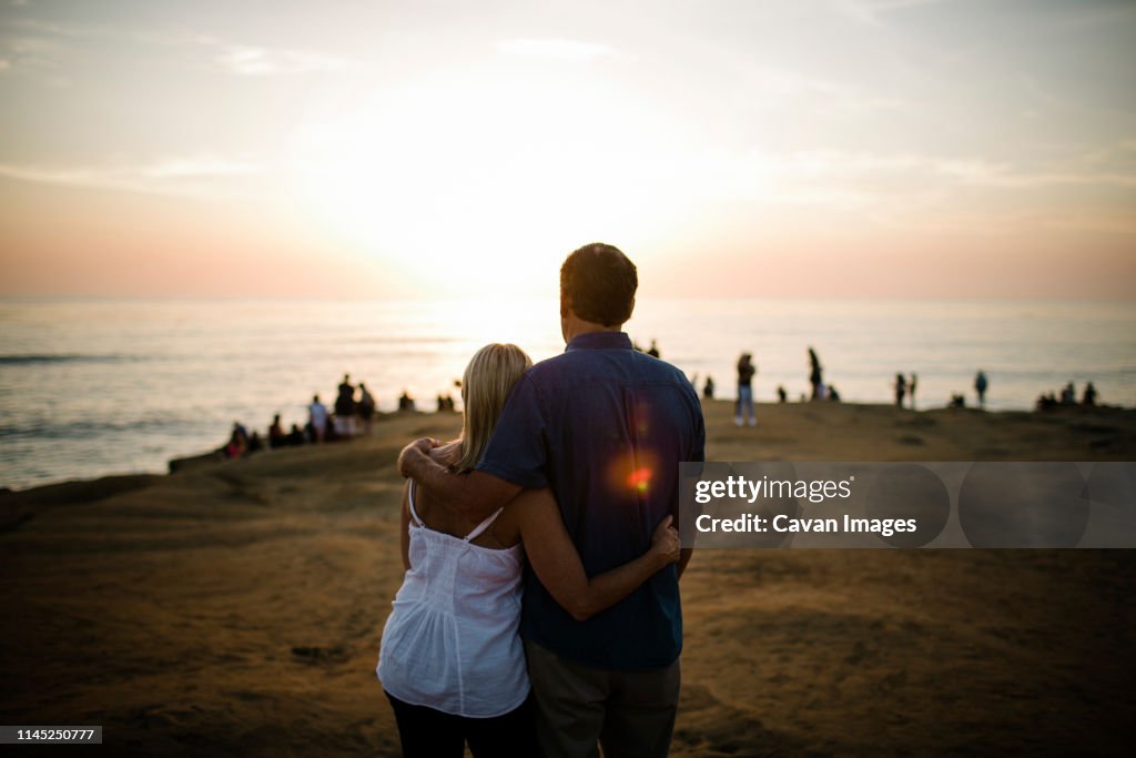Rear view of couple looking at sea while sanding on cliff against sky during sunset