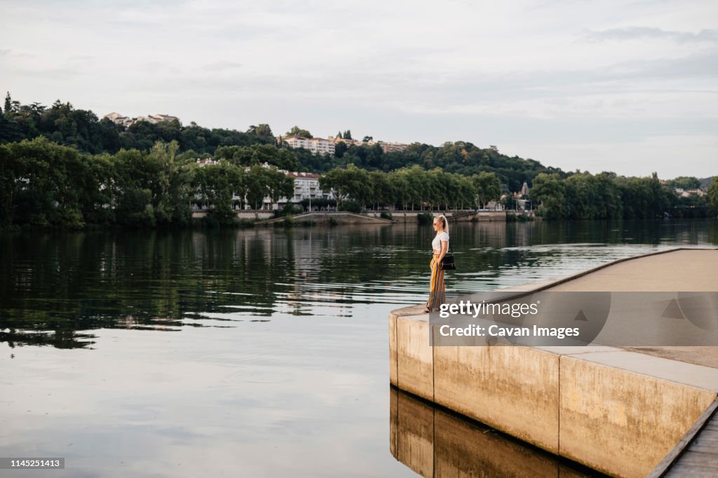 Side view of woman looking at Saone River while standing against cloudy sky during sunset in city