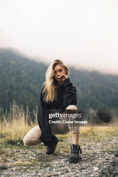 portrait of confident woman crouching on field against mountain in forest during foggy weather - hurken stockfoto's en -beelden