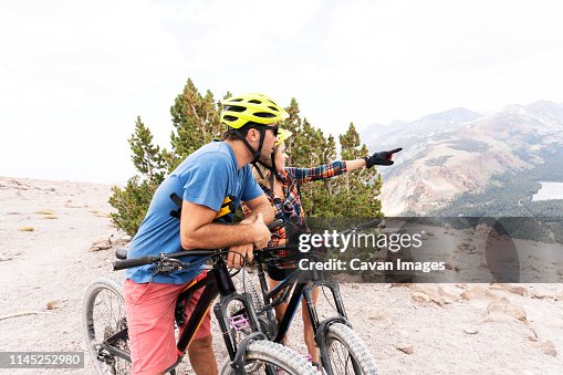 Couple with bicycles looking away while standing on mountain against cloudy sky