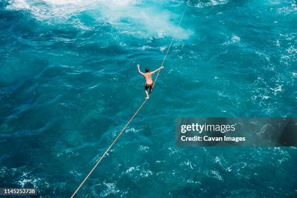 high angle view of carefree young man practicing slacklining over sea - hochseil stock-fotos und bilder