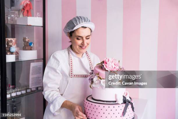 smiling female baker standing by cake against wall in laboratory - confectioner stock pictures, royalty-free photos & images