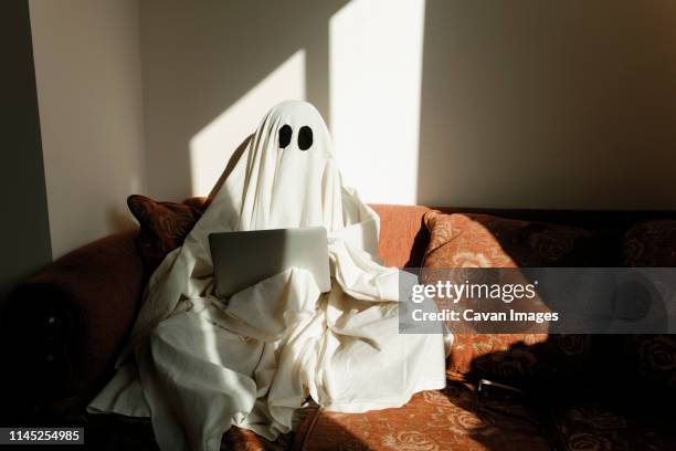 man in ghost costume using laptop computer while sitting on sofa against wall at home - aparición fotografías e imágenes de stock