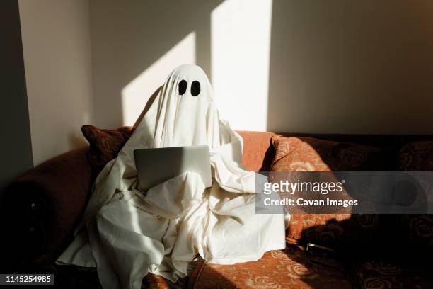 man in ghost costume using laptop computer while sitting on sofa against wall at home - fancy dress costume imagens e fotografias de stock