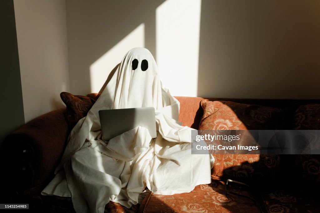 Man in ghost costume using laptop computer while sitting on sofa against wall at home