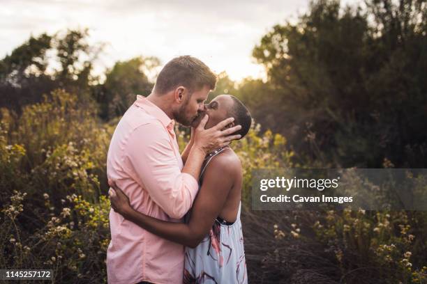 side view of romantic couple kissing while standing amidst plants against sky in park during sunset - beso en la boca fotografías e imágenes de stock