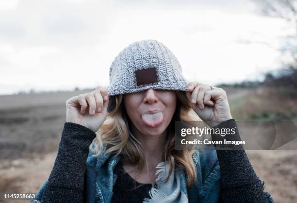 close-up of playful woman covering eyes with knit hat sticking out tongue while standing on landscape against cloudy sky - making a face stock-fotos und bilder