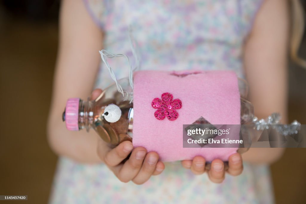 Girl (6-7) holding piggy bank of copper coins, homemade using recycled plastic bottle and craft materials