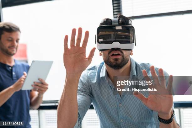jeune homme au bureau expérimentant avec un casque de réalité virtuelle et partenaire à l’arrière-plan contrôlant avec une tablette - casques réalité virtuelle photos et images de collection