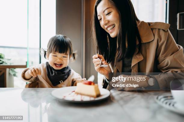 mother and little daughter sharing a piece of cake in the cafe, both of them are smiling joyfully - asian family cafe stockfoto's en -beelden