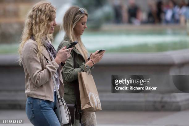 Young women walk cross Trafalgar Square while using their mobile phones, on 20th May 2019, in London, England.