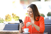 Happy woman using phone stirring coffee in a bar