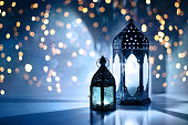 Couple of glowing Moroccan ornamental lanterns on the table. Greeting card, invitation for Muslim holy month Ramadan Kareem. Festive blue night background with glittering golden bokeh lights.