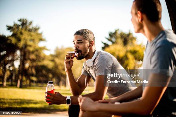aktiver junger mann, der einen biss aus einer proteinstange nimmt und sich neben seinem freund mit einem sportgetränk hydriert - riegel stock-fotos und bilder