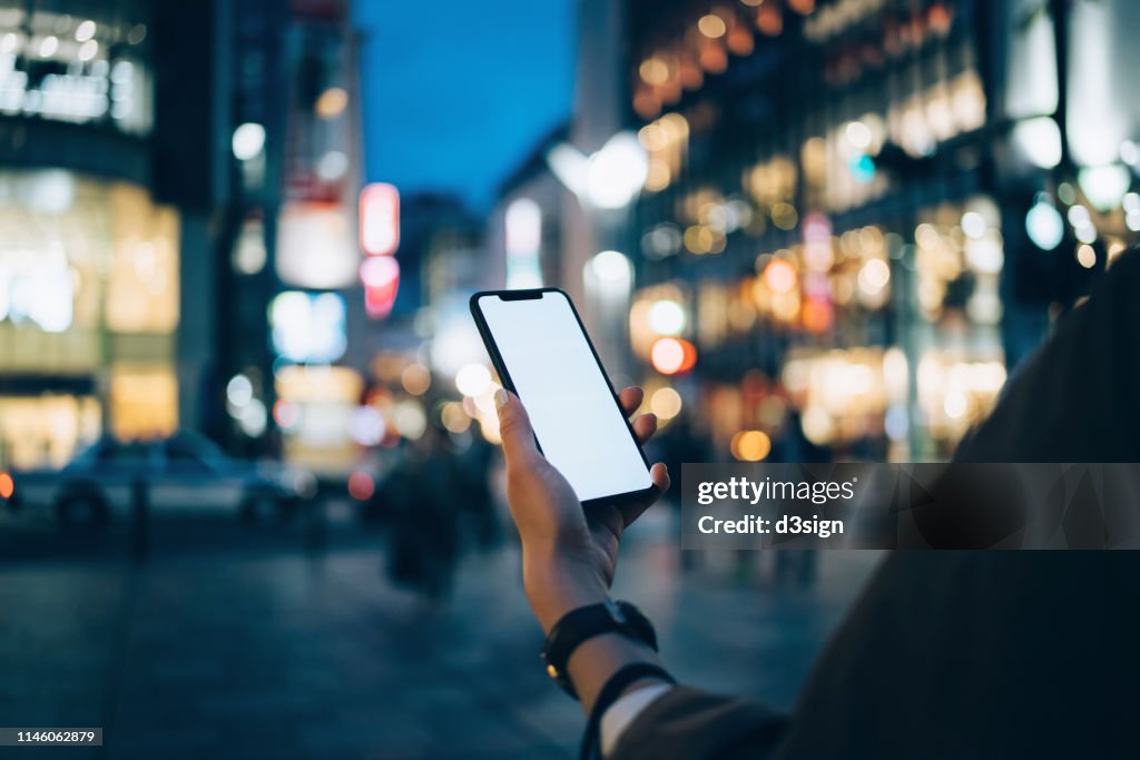 Human hand using mobile phone against illuminated neon commercial sign and city street in downtown district at night