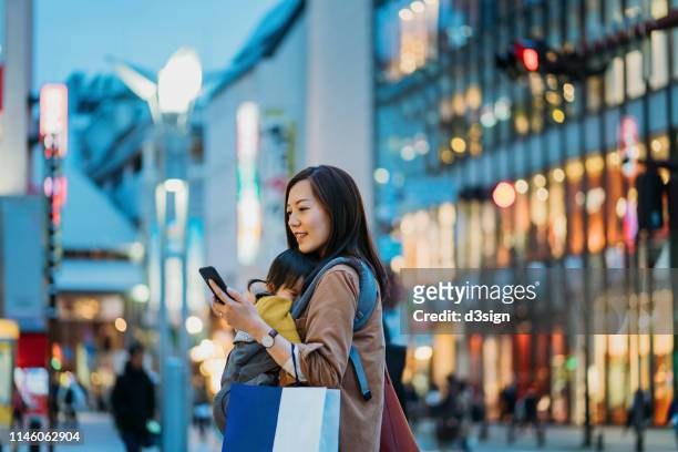 young asian mother carrying shopping bag and using smartphone walking in downtown city street shopping with little daughter - downtown shopping stock pictures, royalty-free photos & images