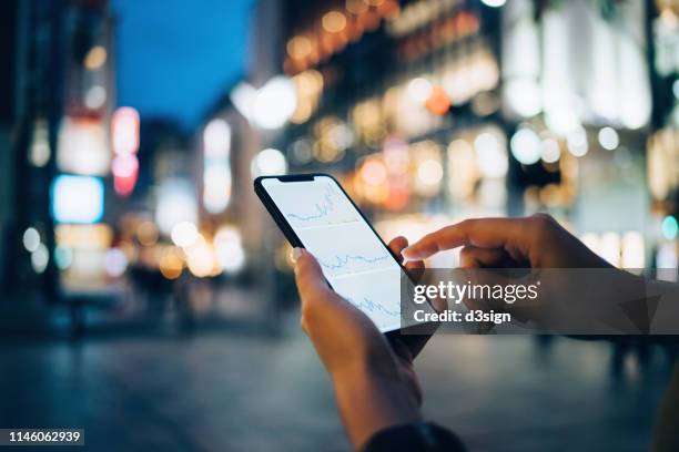 businesswoman reading financial trading data on smartphone in downtown city street against illuminated urban skyscrapers - uomo donna per mano foto e immagini stock