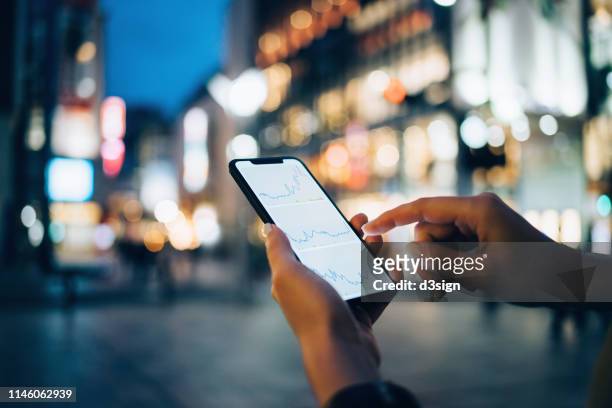 businesswoman reading financial trading data on smartphone in downtown city street against illuminated urban skyscrapers - 電話　ビジネス ストックフォトと画像