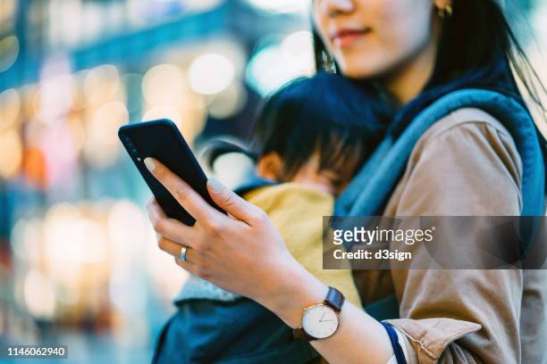close up of young asian mother using smartphone in downtown city street while shopping with little daughter - working mother ストックフォトと画像