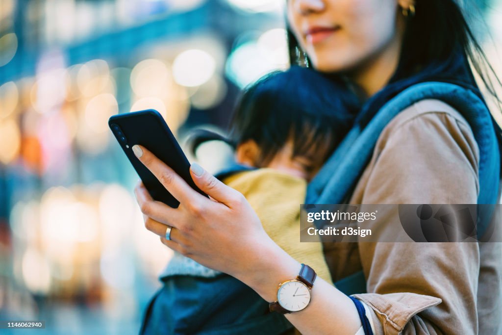 Close up of young Asian mother using smartphone in downtown city street while shopping with little daughter