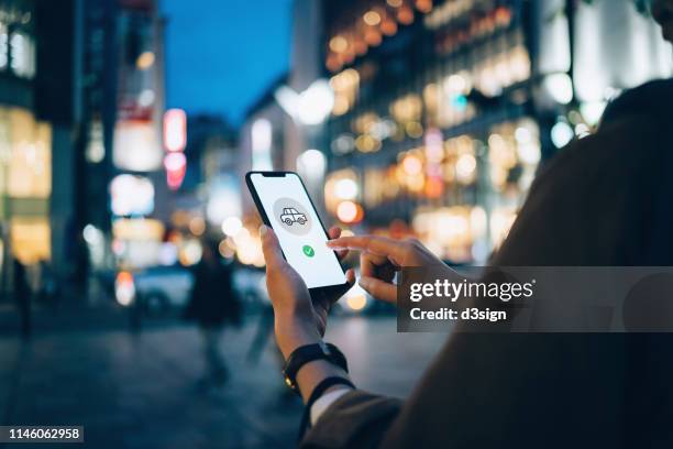 young woman using mobile app on smart phone to arrange taxi ride in downtown city street, with illuminated city traffic scene as background - mobile app car stock pictures, royalty-free photos & images