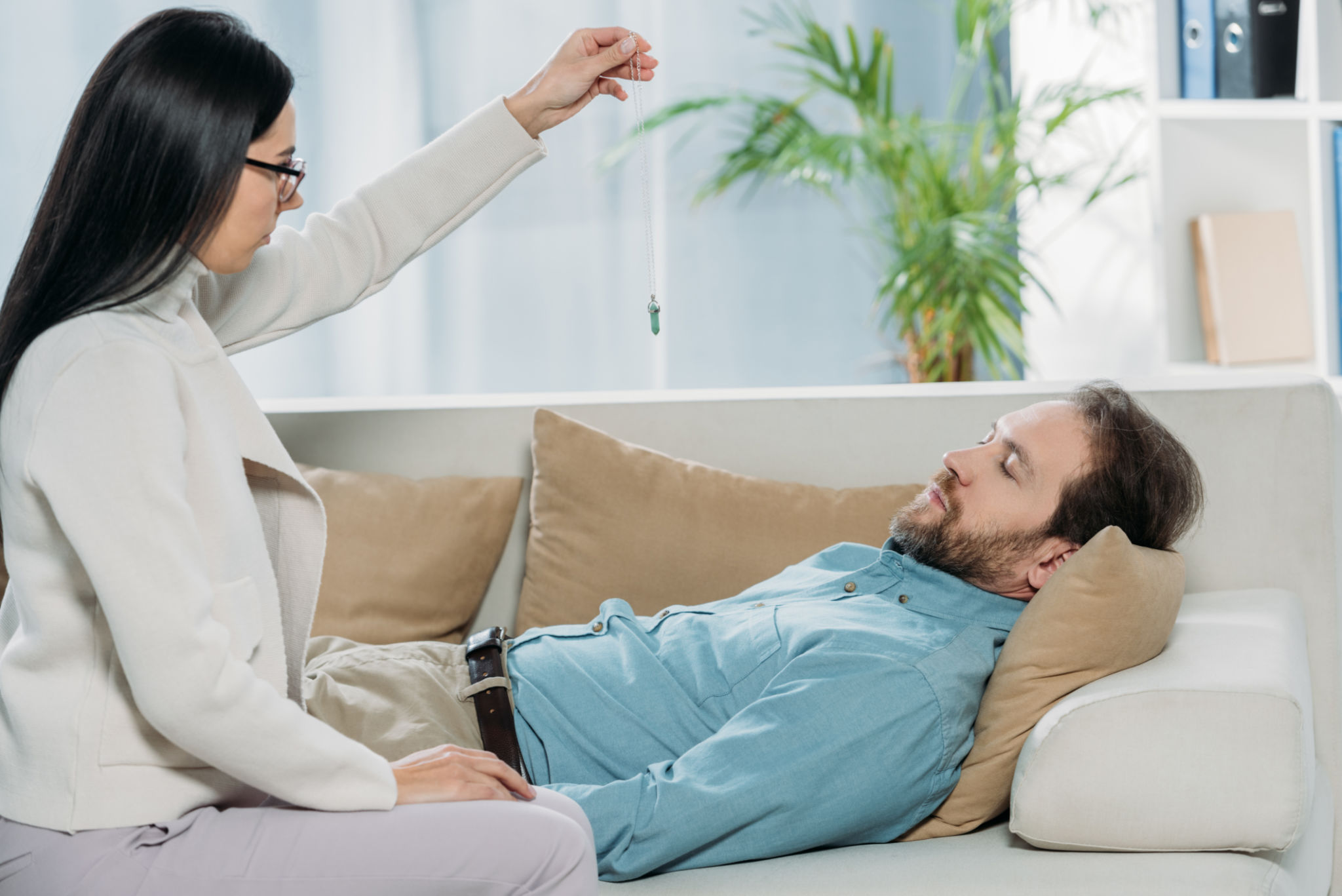 side view of young hypnotist with pendulum and bearded man with closed eyes lying in couch during hypnotherapy side view of young hypnotist with pendulum and bearded man with closed eyes lying in couch during hypnotherapy