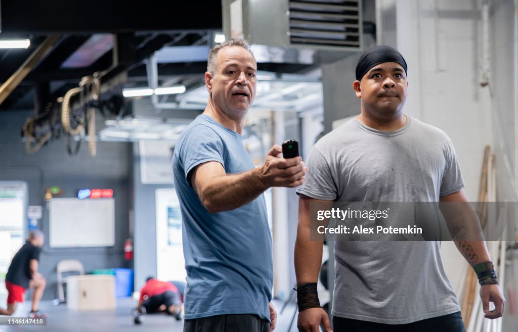 El hombre latino Senior, el entrenador, mostrando los resultados de la aptitud para el joven atleta hispano, levantador de pesas, en un dispositivo de fitness inteligente en el gimnasio durante el entrenamiento.