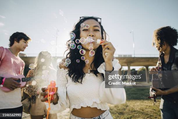 woman blowing soap bubbles while friends playing ukulele during music event - millennials at party photos et images de collection