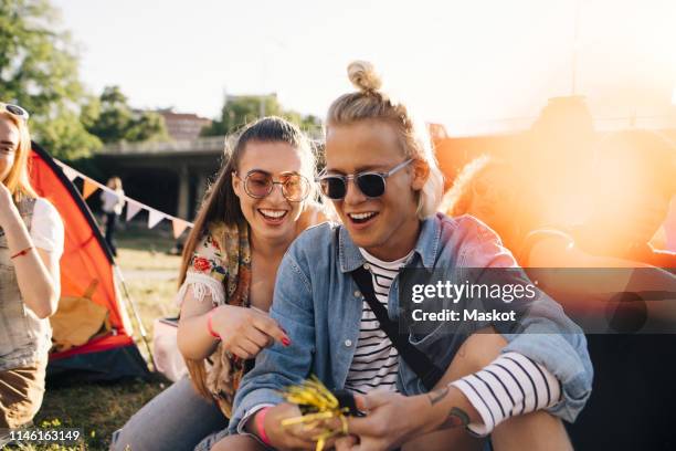 smiling male and female friends looking at smart phone while sitting on lawn in sunny day - hangout festival day 3 stockfoto's en -beelden