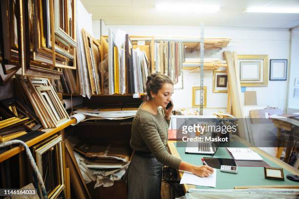 Confident young craftswoman talking on smart phone while writing in paper at workbench in framing store