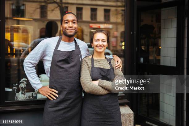 Portrait of confident young owners standing against delicatessen window in city