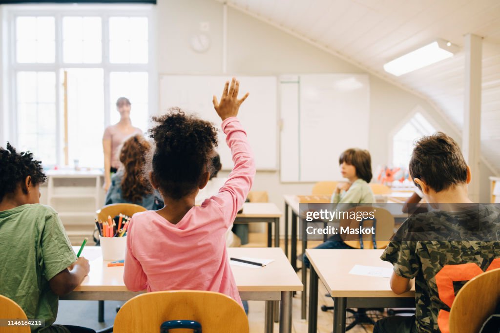 Rear view of boy raising hand while answering in class at elementary school