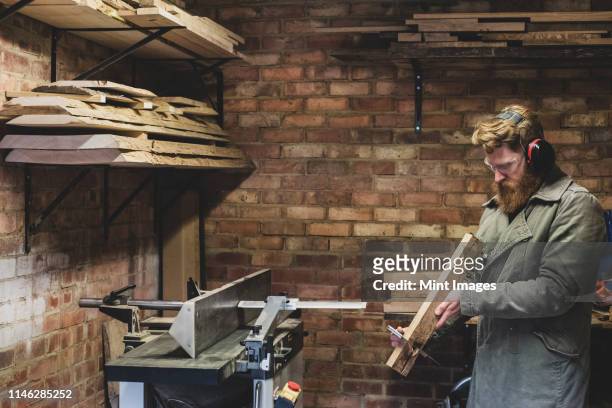 bearded man standing in workshop, wearing ear protectors, working on piece of wood. - traditional parka stock pictures, royalty-free photos & images