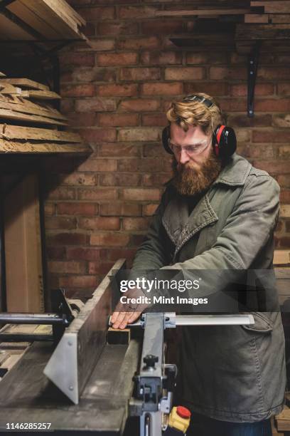 bearded man standing in workshop, wearing ear protectors, working on piece of wood. - traditional parka stock pictures, royalty-free photos & images