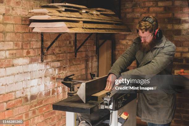 bearded man standing in workshop, wearing ear protectors, working on piece of wood. - traditional parka stock pictures, royalty-free photos & images