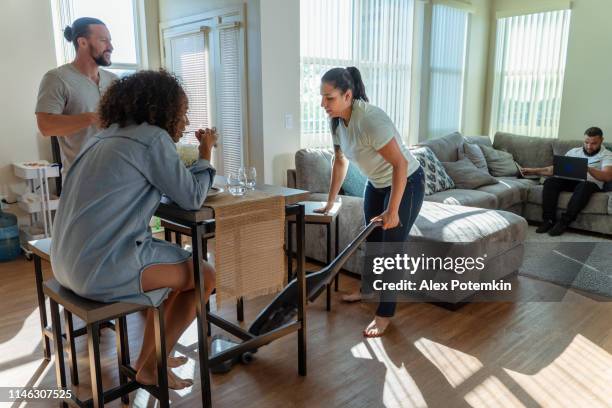die junge hispanic/latino frau, die die gemeinsame wohnung mit einem staubsauger putzt, wenn ihre freunde-mitbewohner haben etwas essen in der küche, und eine andere mitbewohnerin, die auf einer couch in einem wohnzimmer mit pc-laptop. - sharing economy stock-fotos und bilder