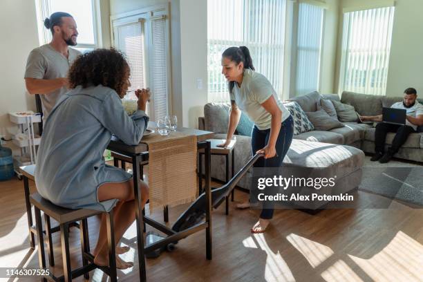 the young hispanic/latino woman cleaning the shared apartment with a vacuum cleaner, when her friends - roommates have some food in the kitchen, and another roommate working in on a couch in a living room with pc laptop. - friends clean stock pictures, royalty-free photos & images