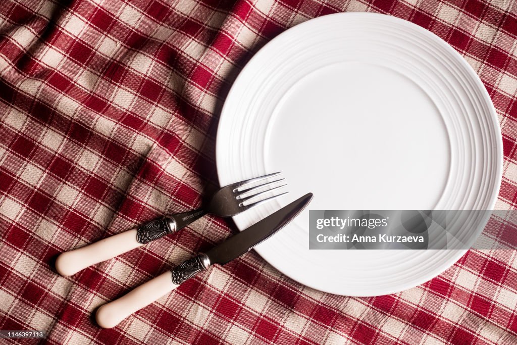 Empty white plate, fork and knife on a red and white linen checkered napkin, top view. Image with copy space. Kitchen table with a towel and a plate - top view with copy space.