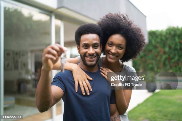 young couple in front of a house, holding keys of their new home - celebrates firsts imagens e fotografias de stock