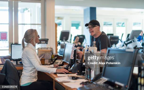 the senior man asking for information about the gym and having a conversation with the senior silver-haired woman who working at the reception. - health club desk stock pictures, royalty-free photos & images