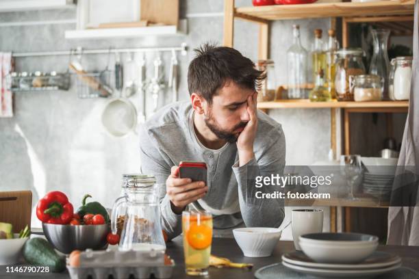 hombre triste usando teléfono inteligente durante el desayuno en casa - disgusto fotografías e imágenes de stock
