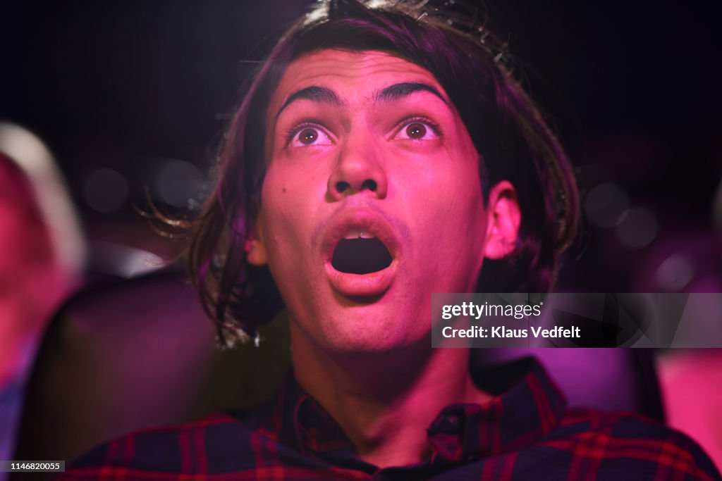 Surprised young man with mouth open watching horror movie in cinema hall at theater
