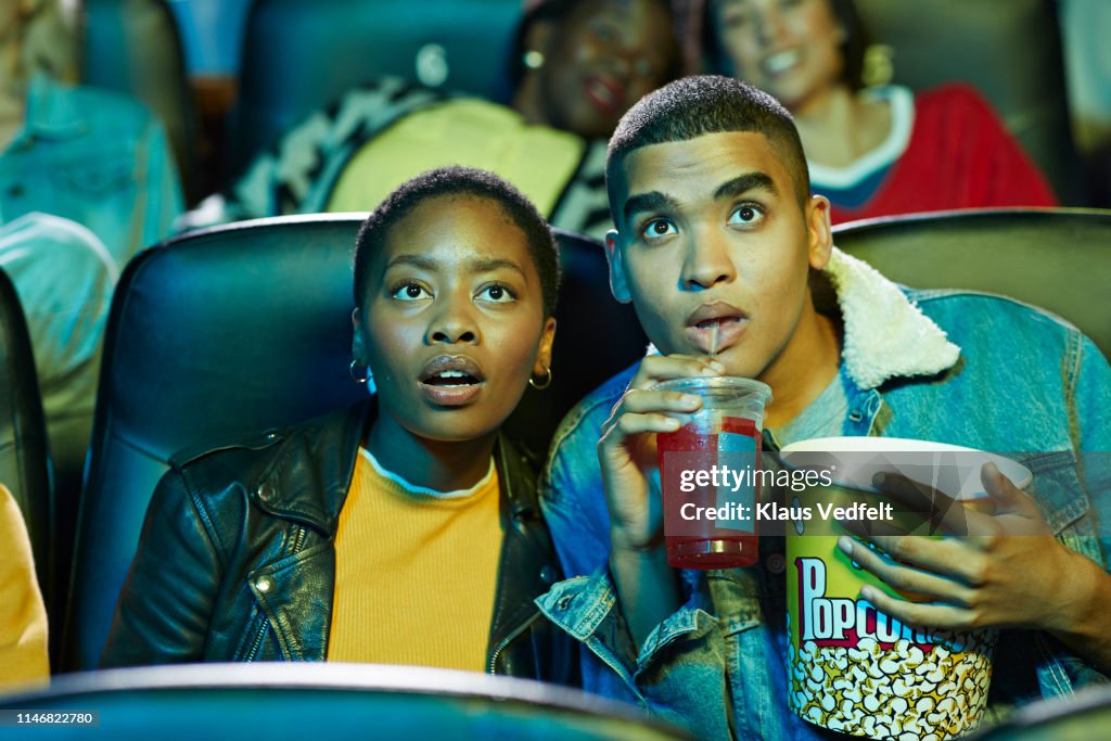 Surprised young man drinking soda while watching movie with friend in cinema hall